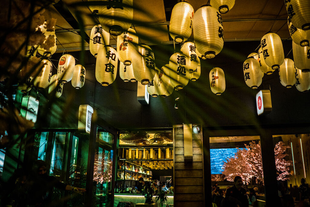 The exterior of the Paperfish Sushi Bar terrace at night with white paper lanterns and Japanese characters.